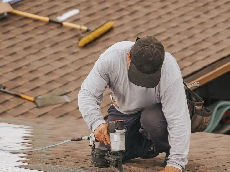 A roofer kneeling on a shingled roof, nailing a course of shingles down.