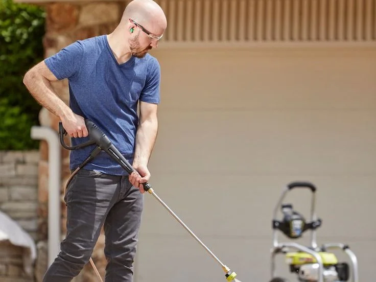 A man washing a driveway with a pressure washer, the clean strip showing behind the jet.